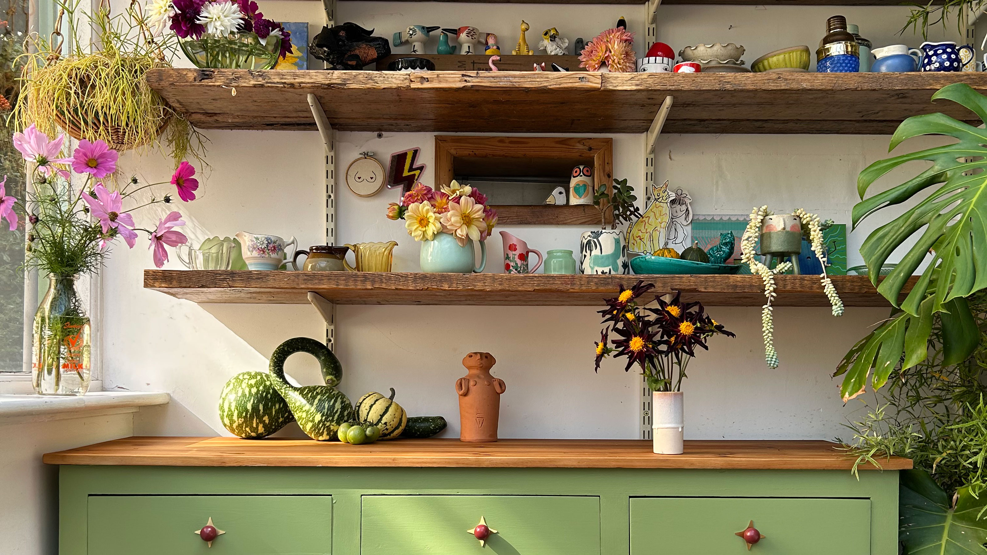 Green kitchen cabinets below open wooden shelving full of vegetables, mugs and vases of flowers