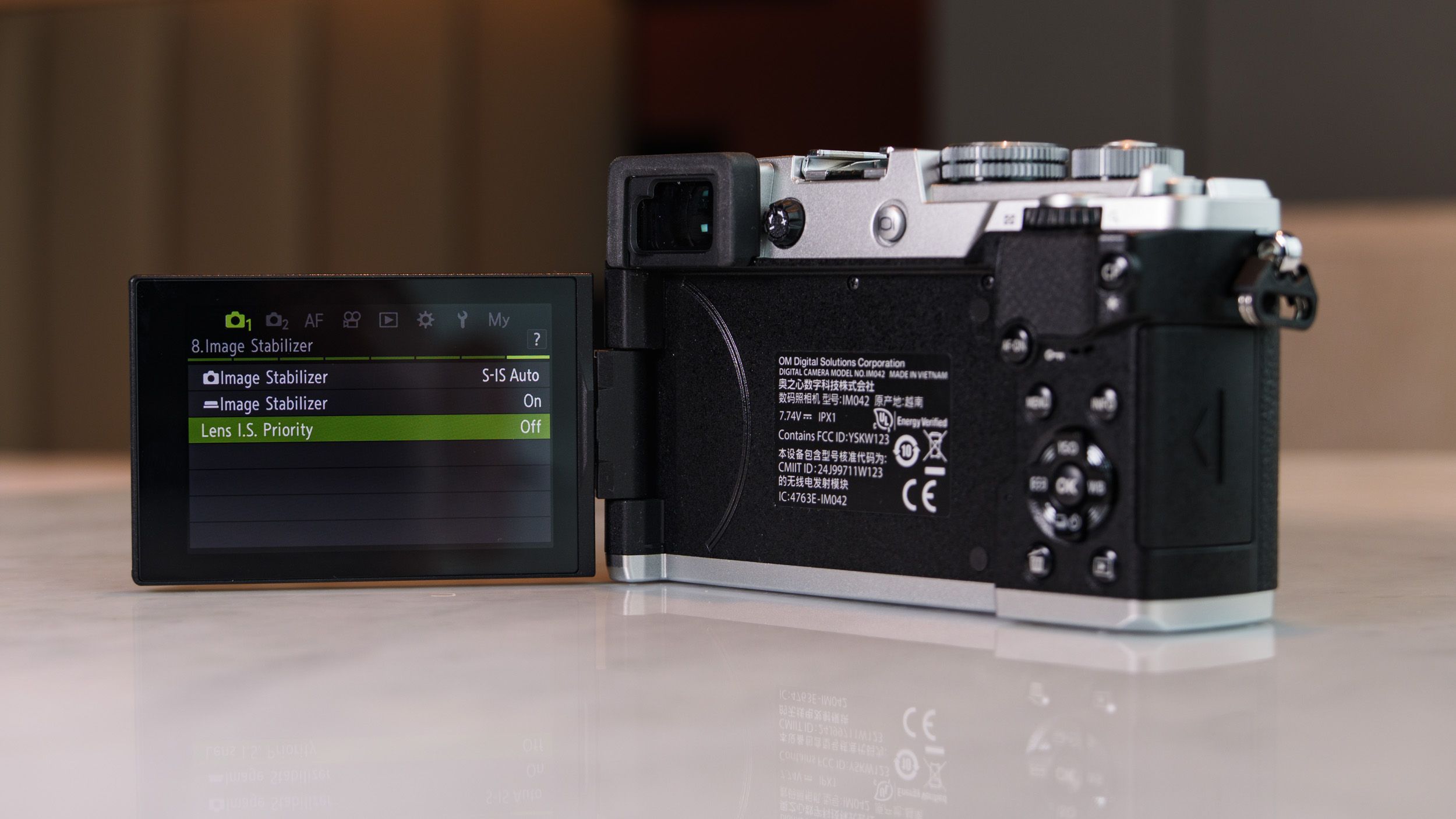 Rear view of the OM System PEN mirrorless camera with its screen flipped out to the side on a white marble surface in a modern room with lantern in the background