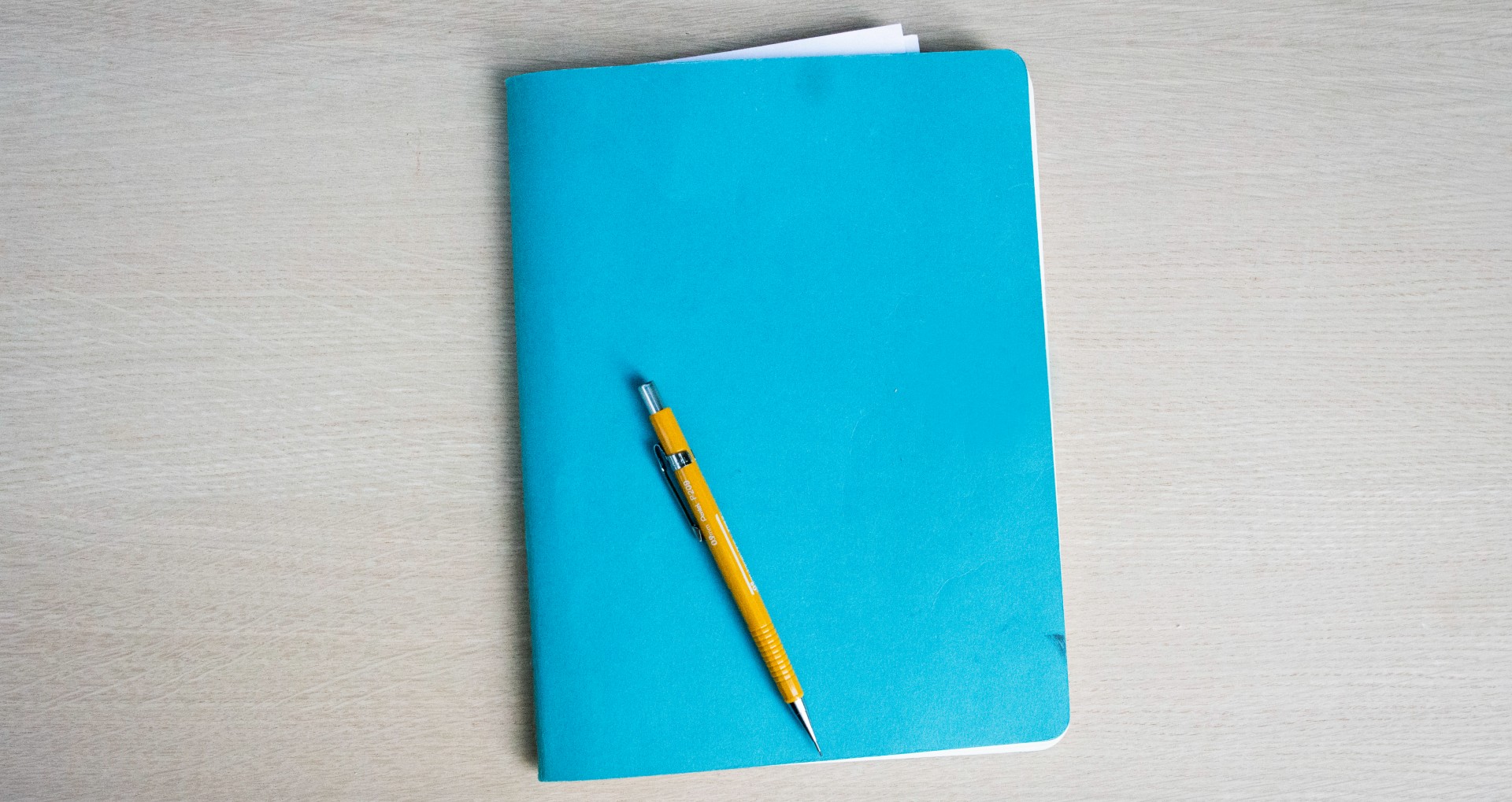 plain, bright teal notebook with a yellow mechanical pencil resting diagonally on top of it, set on a light-colored wood surface.