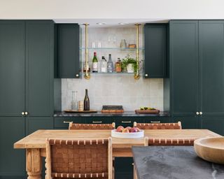 A kitchen with dark green cabinets, a glass and brass shelf and a farmhouse-style wooden dining table