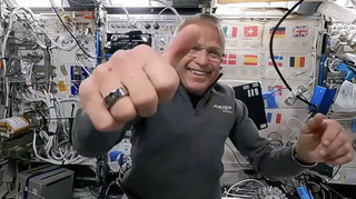 an astronaut gives a thumbs up to the camera in space. the right hand is very close to the camera and a ring is visible on the index finger. behind, the astronaut smiles widely while wearing a shirt that says "axiom space." the background shows a module with flags and various wires and pieces of equipment