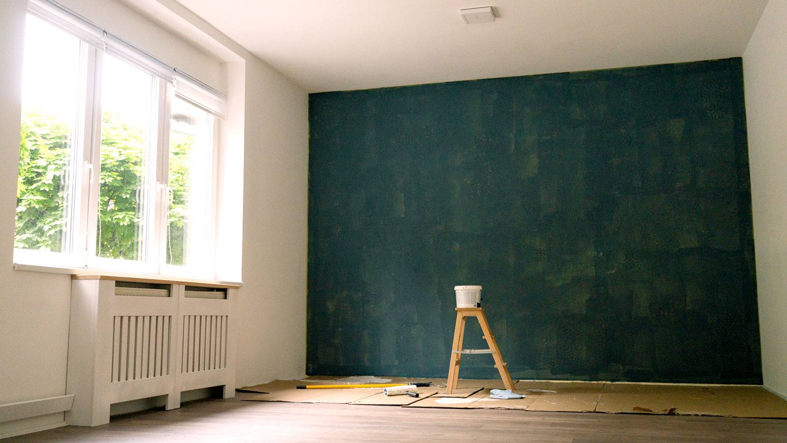 White room with large window on left and dark painted feature wall with step ladder, paint pot and dust sheets on floor