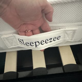 A hand holding the side handle on the Sleepeezee Jessica mattress. The word 'Sleepeezee' is embroidered on the handle in navy cotton