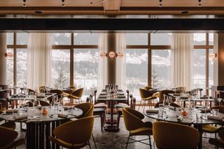 A playfully furnished ski hotel's restaurant features velvety seats, circular Art Deco lighting, and terrazzo floors, while its floor-to-ceiling windows let snowy views in.