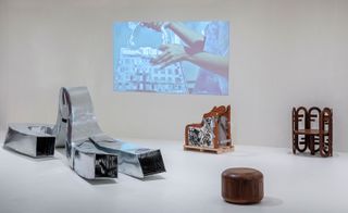 Installation view of Mabeo and Fendi's collection at Design Miami featuring a silver air duct style sculpture, a chair prototype, a wooden chair, a wooden stool and an image on the wall of a person holding a handbag. The pieces are on display in a room with a light grey floor and walls