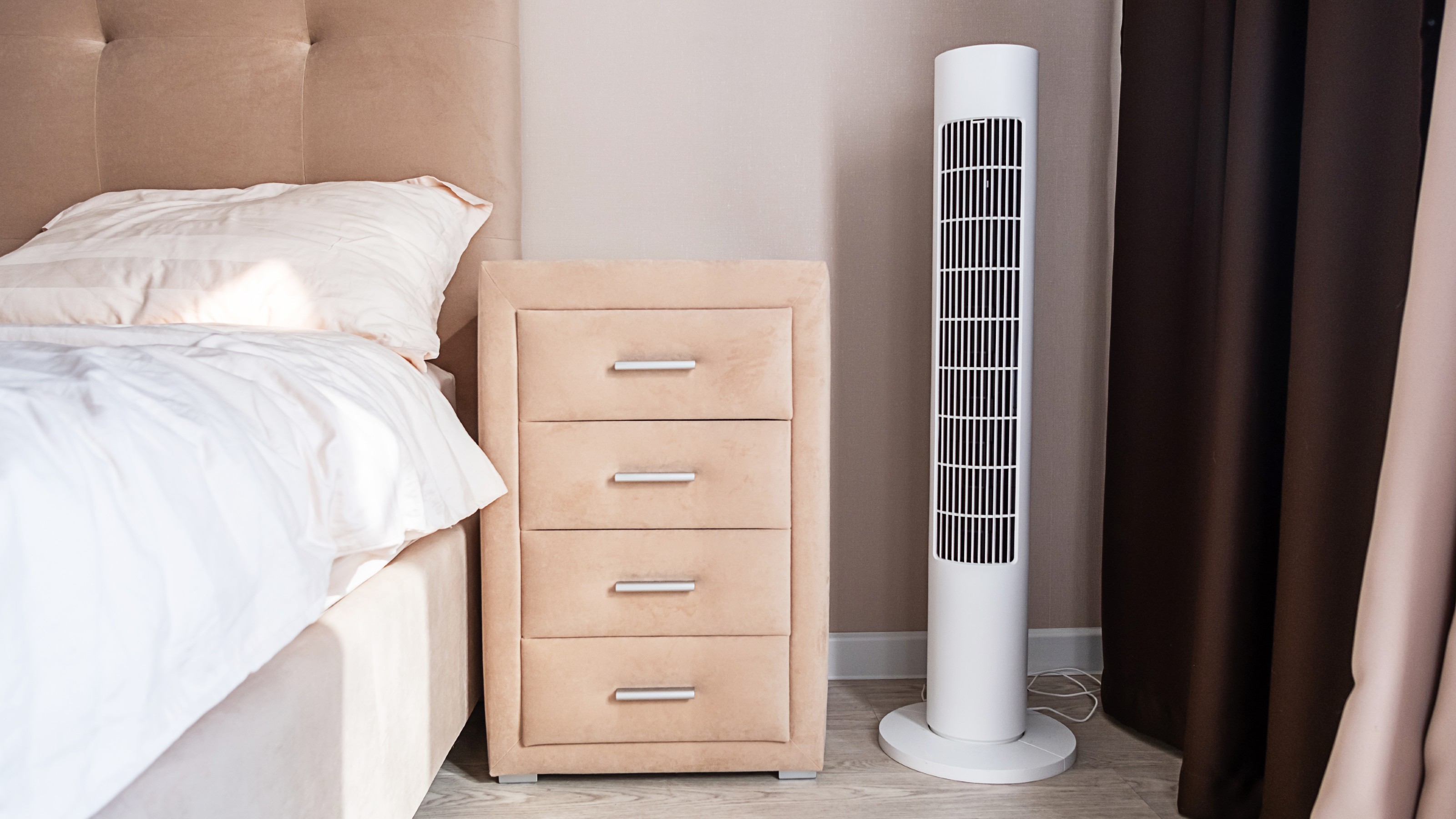 White tower fan on the floor near a bedside table next to a bed with a padded headboard
