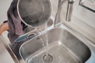 A woman pouring boiling water from a pan down a kitchen sink