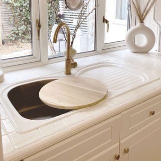 Silver steel sink dropped in white tile kitchen counter top with a gold tap and wooden cutting board on sink