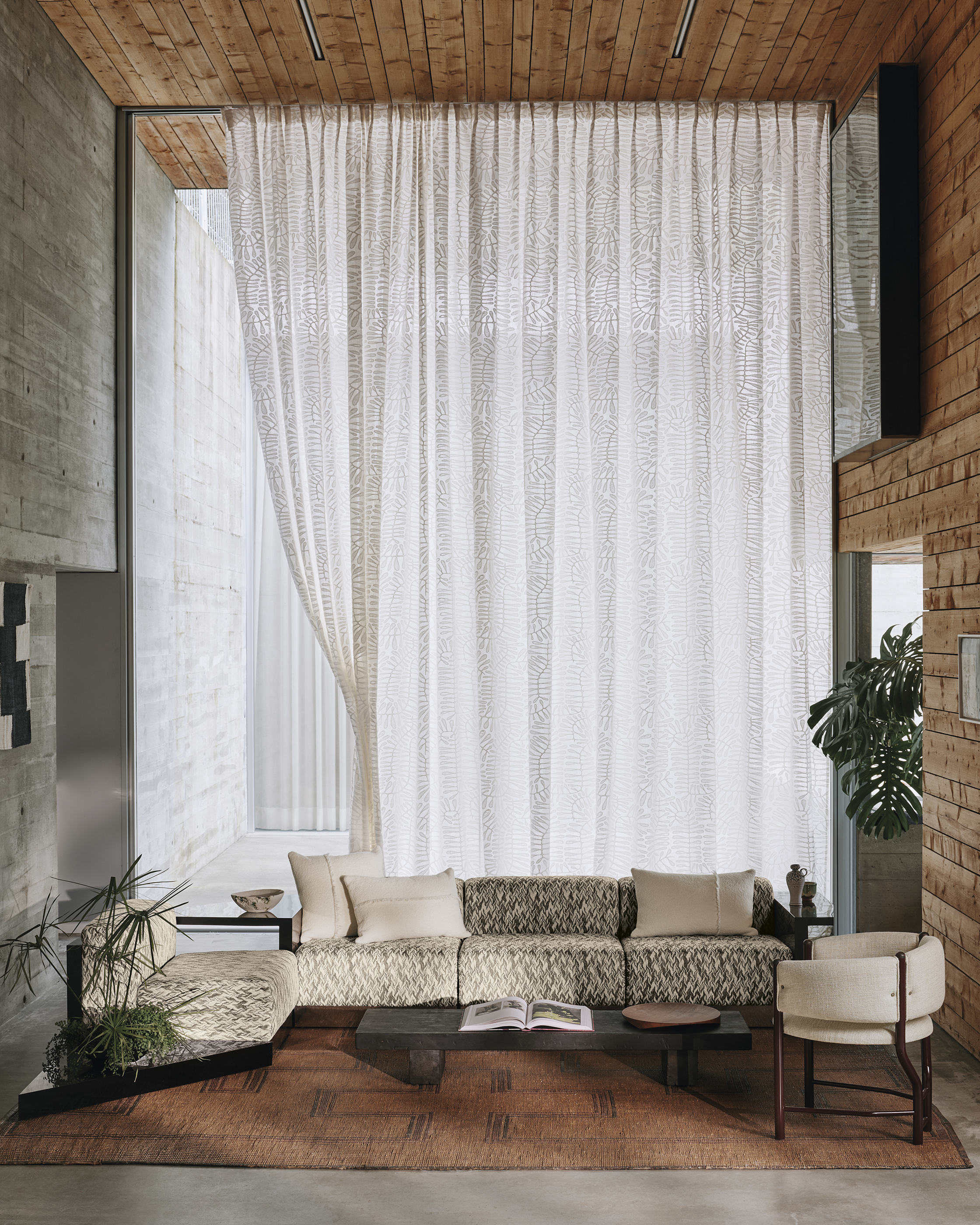 large modern living room with double-height ceilings and wall of sheer curtains, timber paneling, rust-colored rug with patterned sofa and white cushions, and a concrete wall