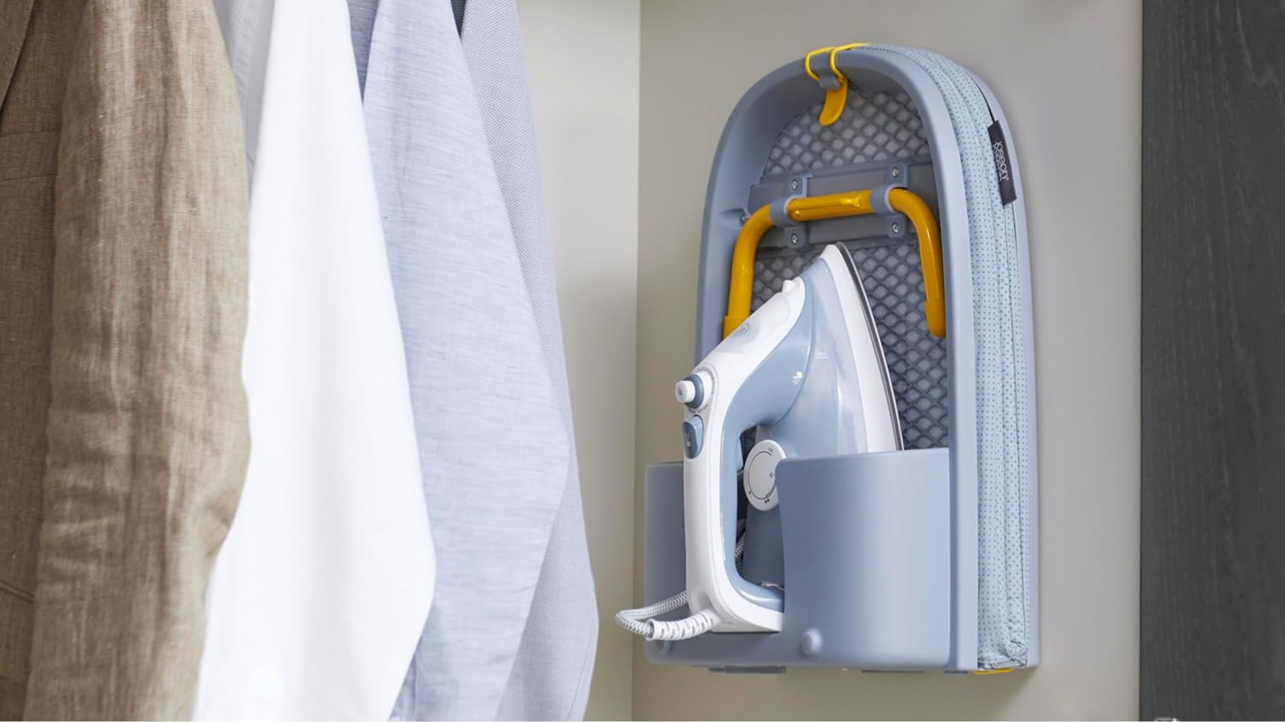 Joseph Joseph compact ironing board hanging in an open wardrobe next to beige, white, and blue garments.