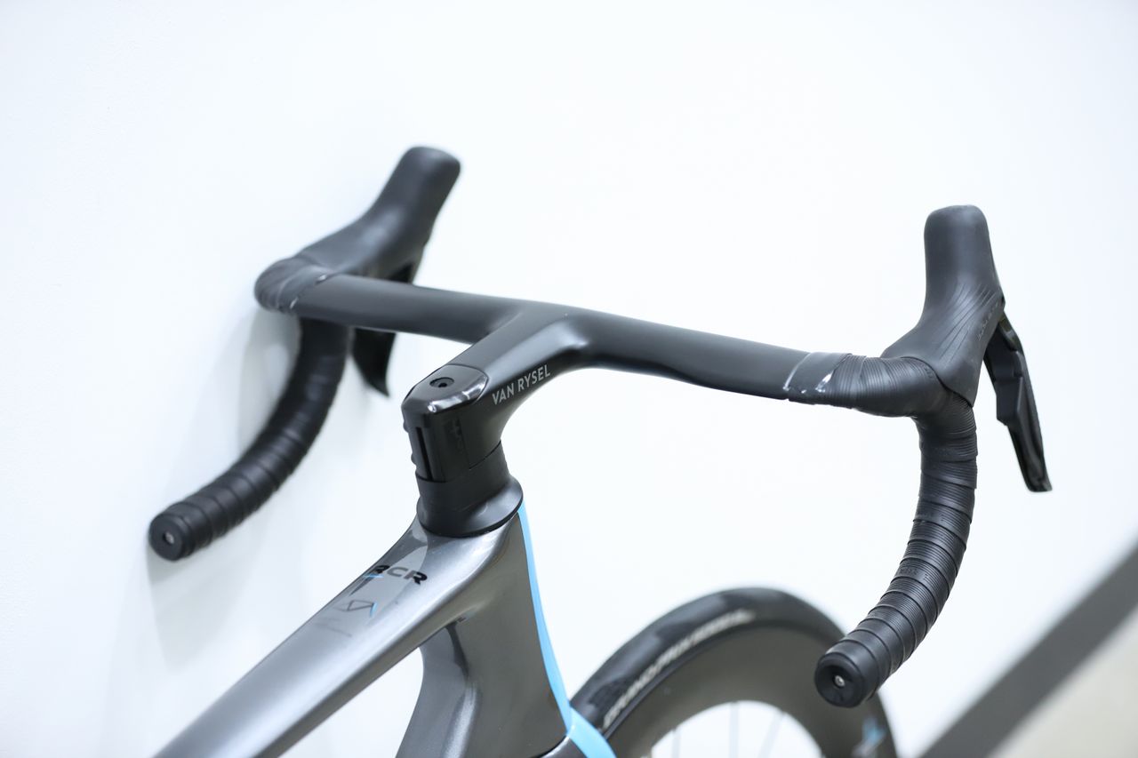 A blue and grey Van Rysel RCR Pro stands against a white wall
