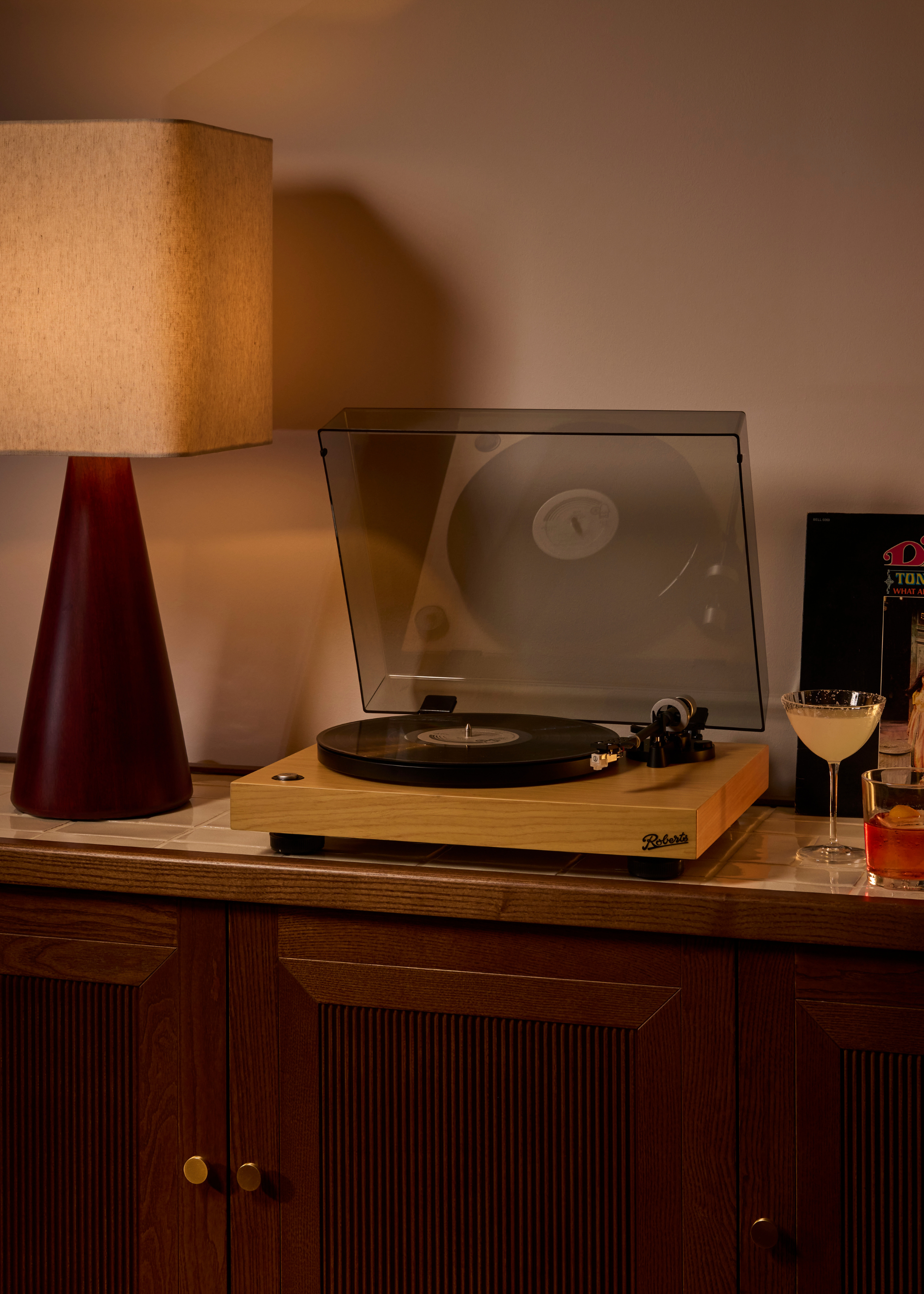 A clos-up of a living room corner with a wood sideboard holding a table lamp, a record player, cocktails and a record