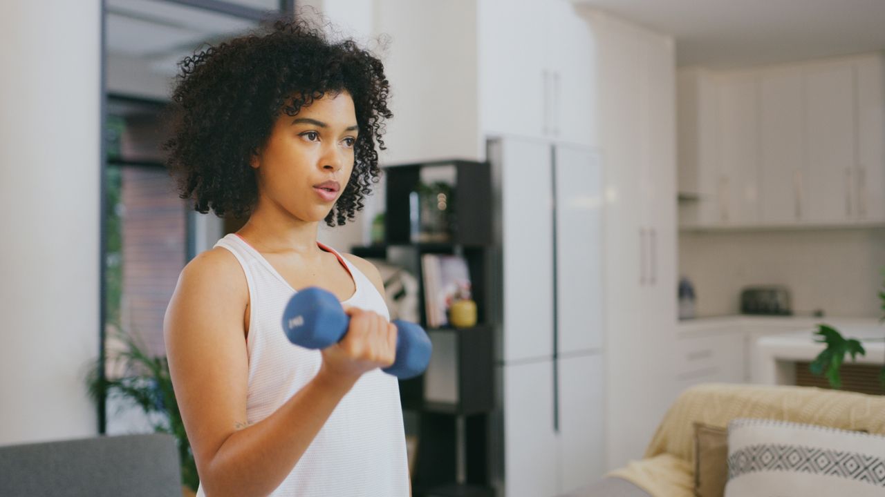 woman in a kitchen home setting wearing a white vest and holding a dumbbell in a biceps curl position. 