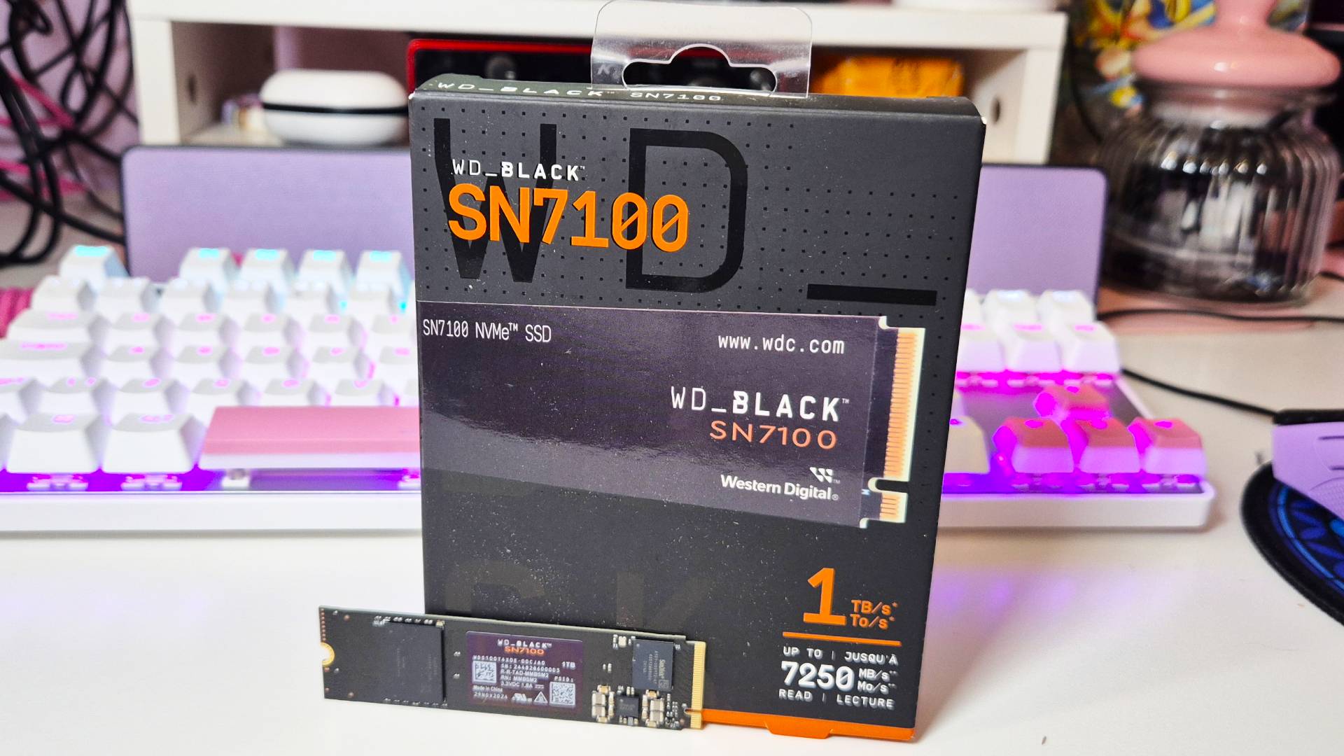 Image of a black SSD sitting in front of a black and orange box, with an RGB keyboard sitting behind both.