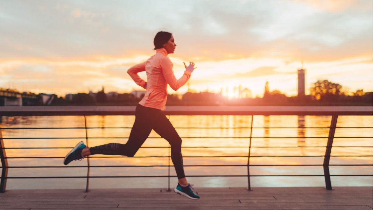 A woman runs alongside a river, with a city scape in the background. She is wearing leggings, sneakers and a long-sleeved top