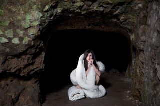 A woman wrapped in a huge knitted scarf is kneeling in a cave-like space.