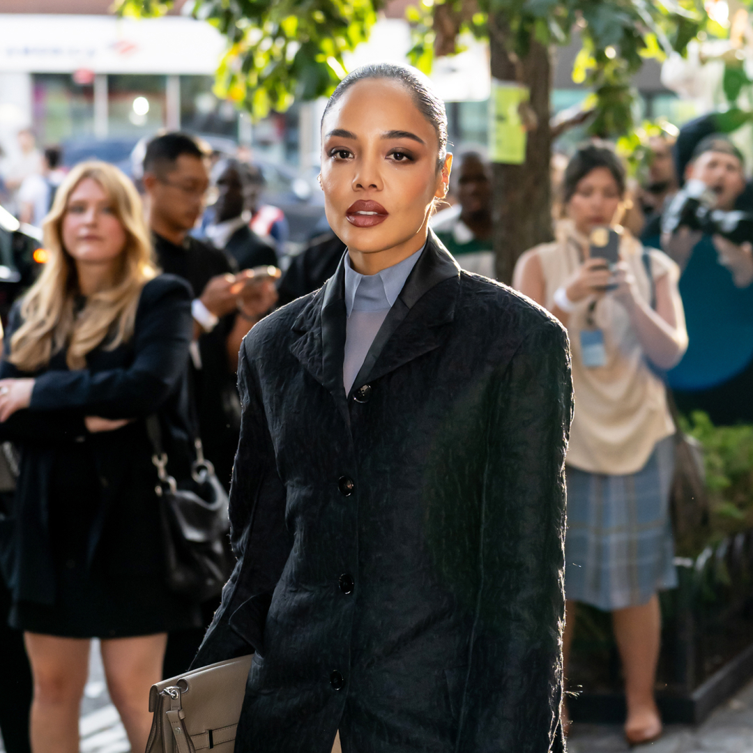 Tessa Thompson wearing a slicked-back bun hairstyle, a blazer, a button up shirt and dark pink lipstick. 