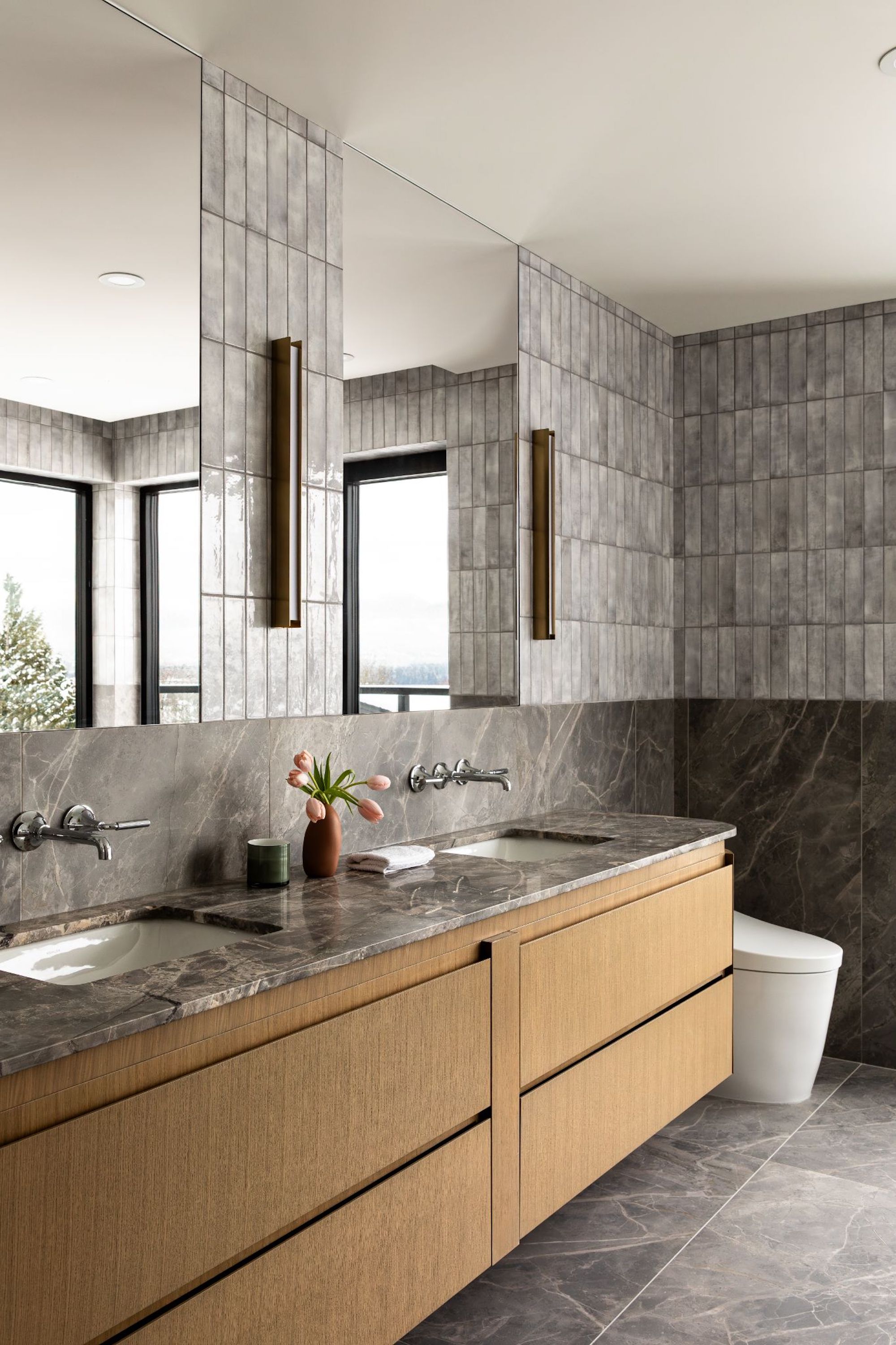 A view of the bathroom sink area, with grey marble work tops and floor tiles, two tall mirrors are fitted above both sinks and vertical lights are fitted in between.