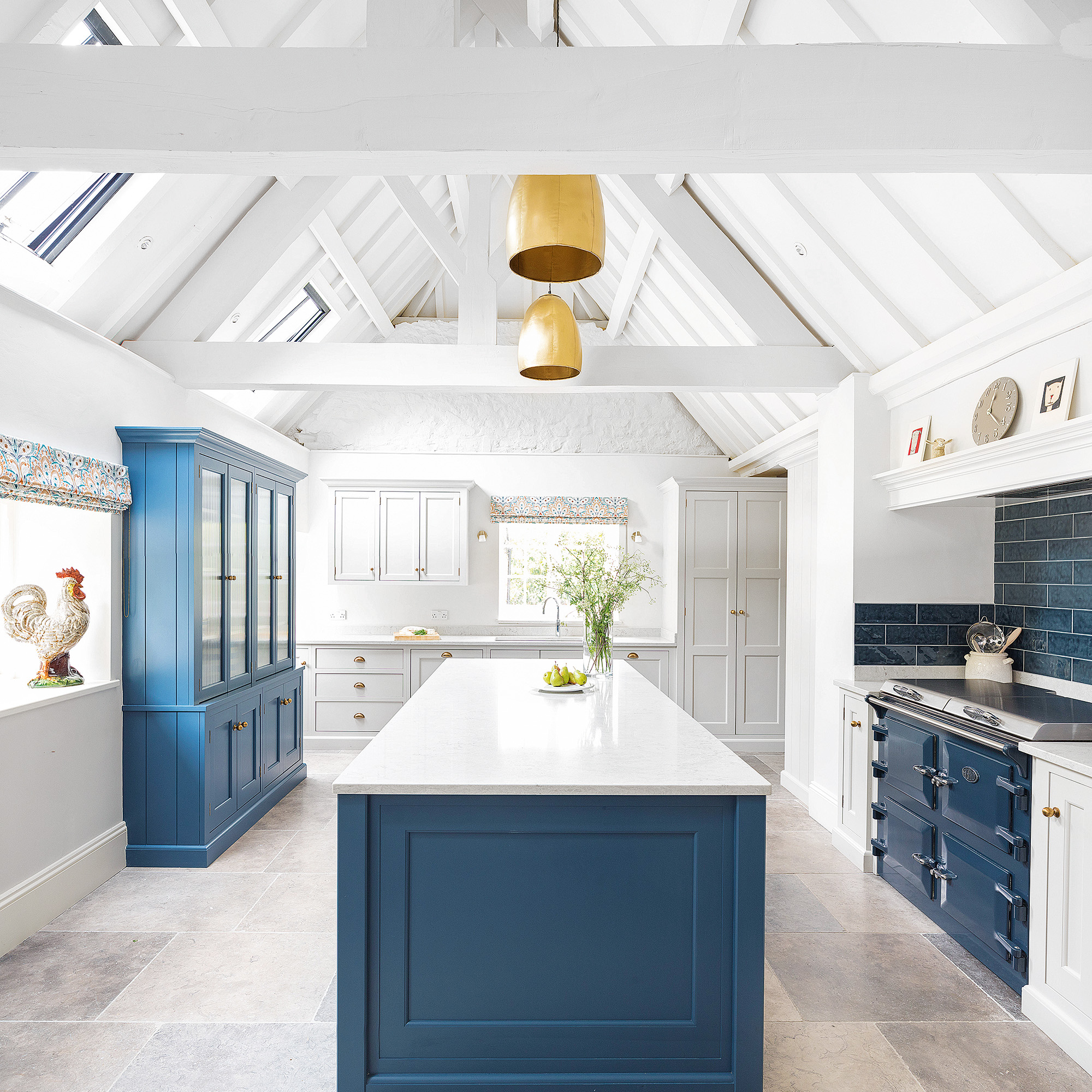 kitchen with white walls, white vaulted ceiling and white cabinetry with a blue kitchen island, blue range cooker and dark blue metro tiled splashback two brass pendant lights in centre with grey stone flooring