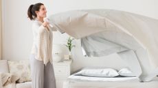 A woman stands next to her bed as she shakes her duvet into the air