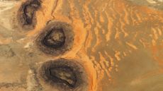 An astronaut photo of three dark mesas, partially surrounded by orange sand dunes