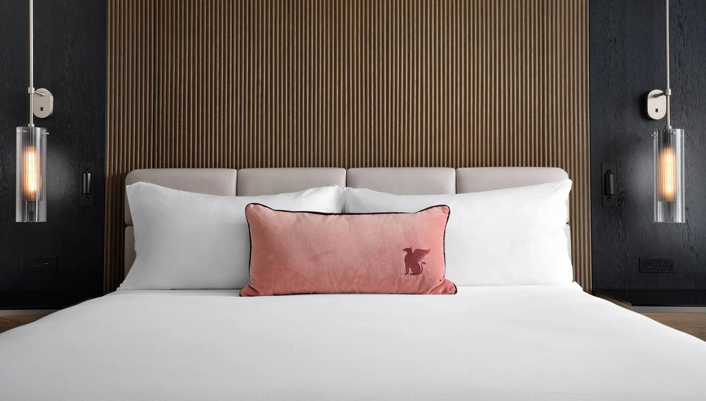 A close up of a Marriott Bed in a JW Marriott Hotel room, showing a bed well made with white sheets, two pillows propped against a quilted headboard, and a pink cushion with the JW Marriott logo on it resting in front of the pillows