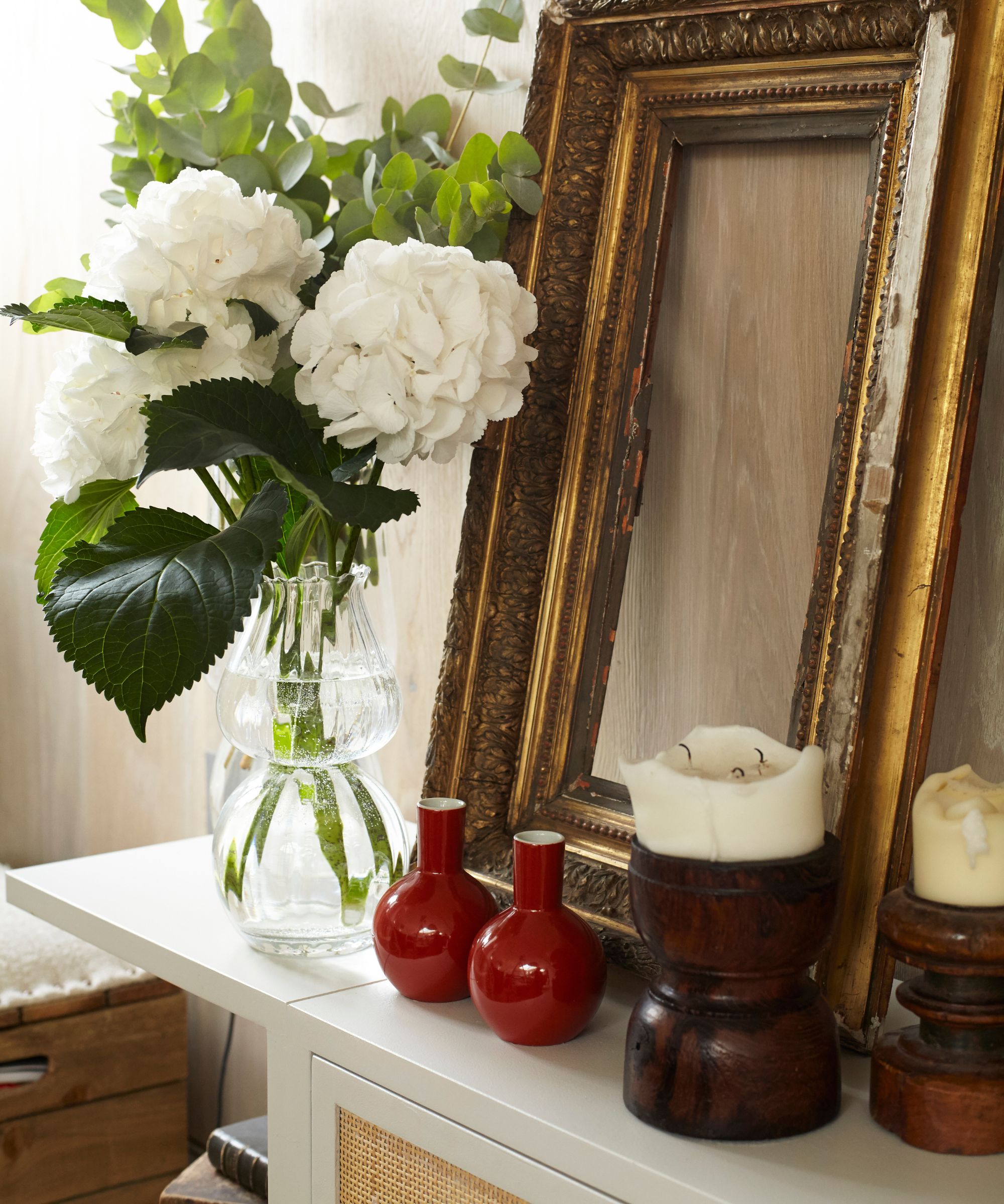 Candles with an empty gold frame, white flowers in a clear vase, two small red vases, and two wooden candle holders with white pillar candles.