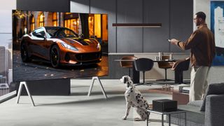 A large LG M5 OLED TV sitting in the corner of a posh high-rise condominium. There's an image of a red sports car on the screen and it's being watched by a man and a dalmatian dog.