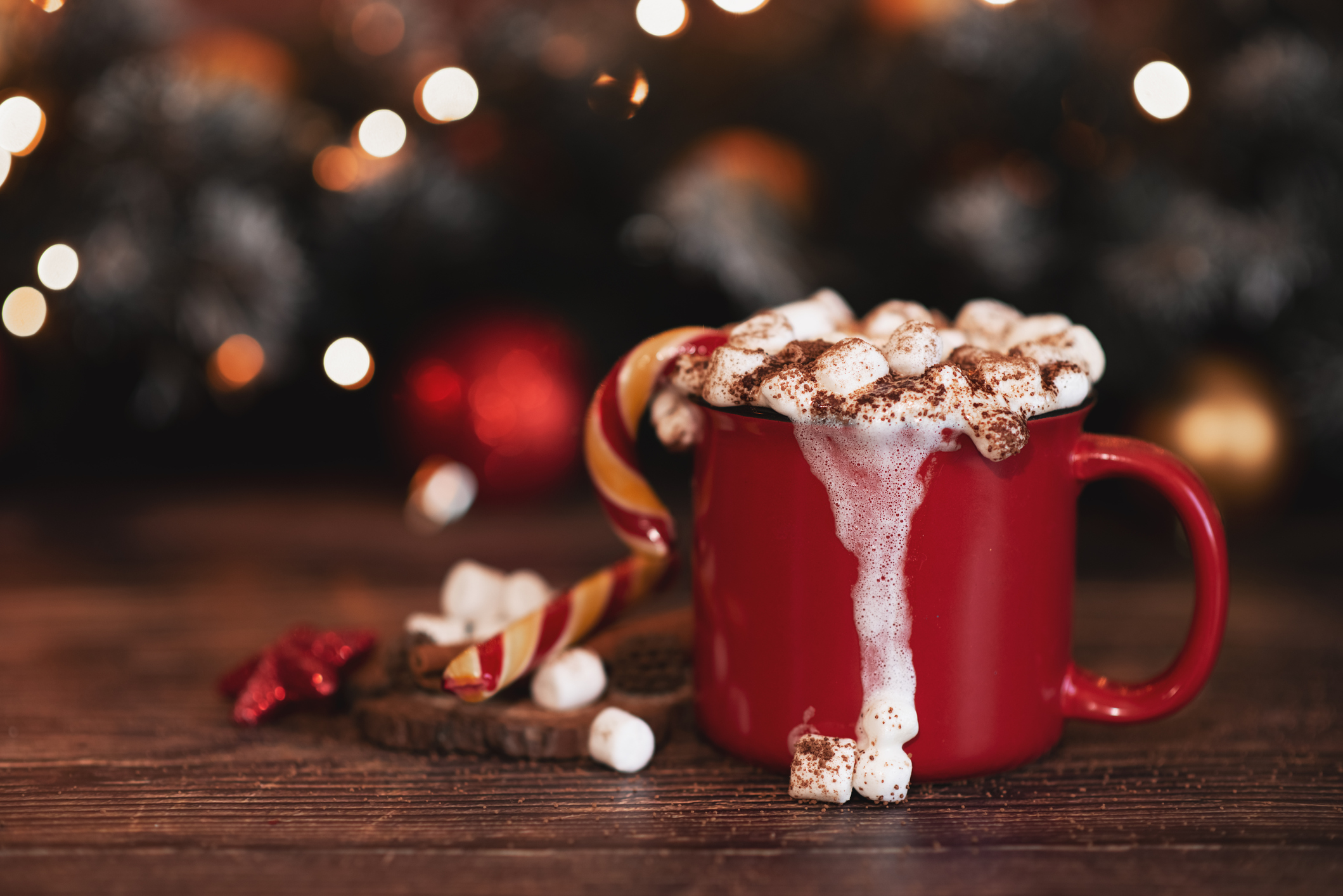 Hot chocolate in a red mug with marshmallows on top placed on a wooden table with Christmas lights behind it.