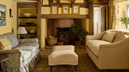 cozy country cottage living room with butter yellow walls, a brick fireplace, beams, a blue pattern skirted sofa with a yellow check pillow and a patterned ottoman