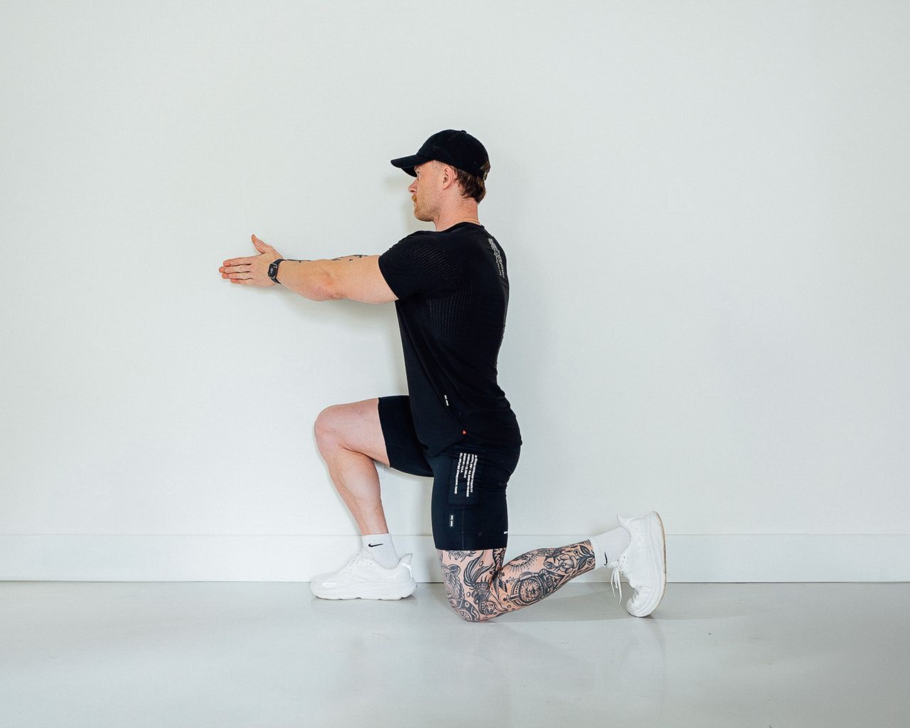 Coach Corey Perkins performs a half-kneeling stretch. His left leg is bent at a 90-degree angle, with his knee and toes resting on the floor. His right leg is also at a 90-degree angle, but his right knee is up in line with his hip and the sole of his right foot rests on the floor. His torso points in the same direction as his knees and his arms are held straight out in front of him with his palms resting together.