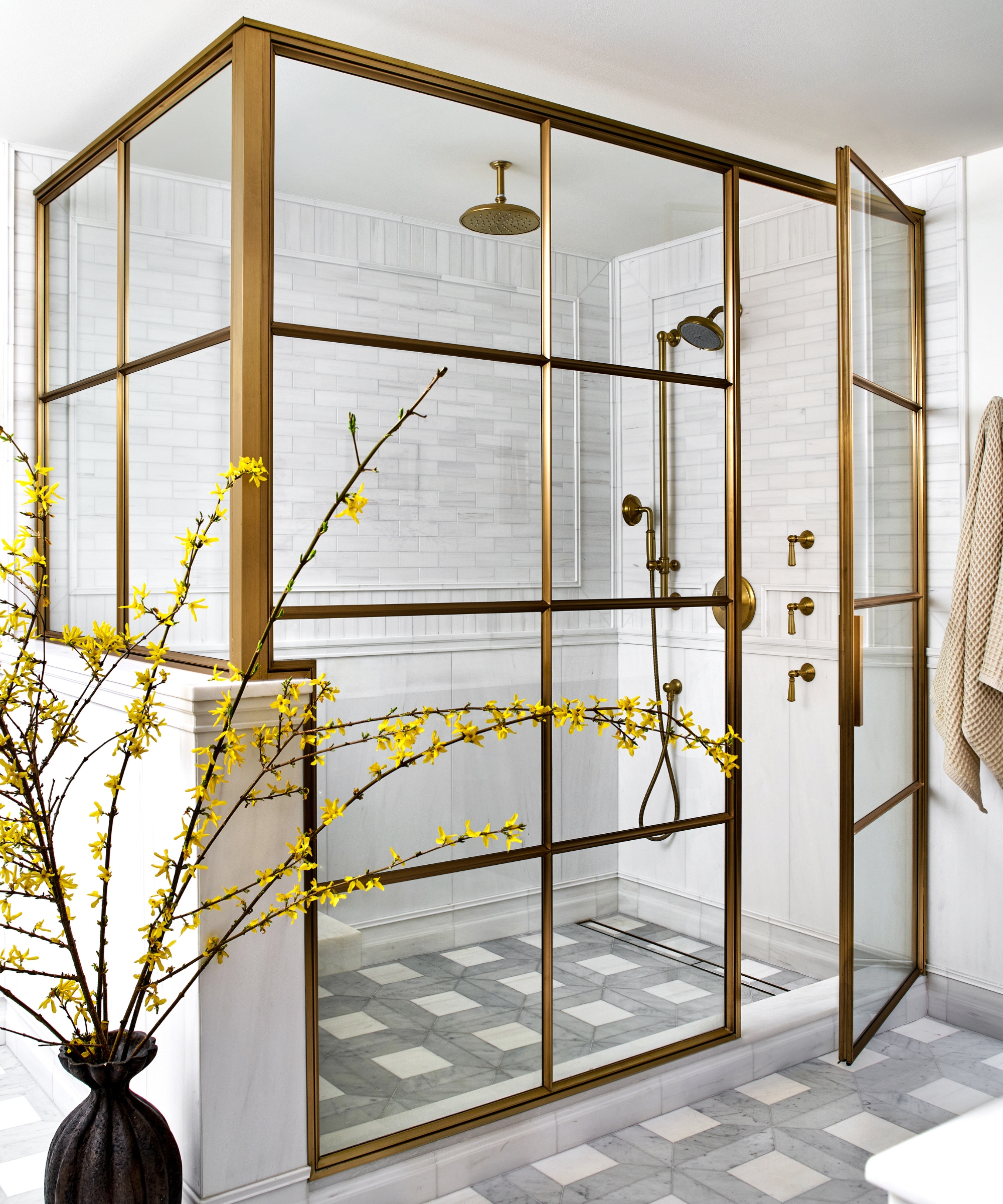A neutral bathroom with a large walk in shower surrounded by a glass and brass enclosure, white wall tiles, and a unique gray and white geometric floor tile layout