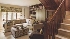 Small neutral living room with ornate wooden staircase and built in cabinetry at the far back wall. There is a patterned square coffee table in front of an L-shape sofa. It looks cozy but bright