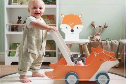 A smiling toddler stands while holding the handle of the GLTC Wooden Fox Walker, one of the best baby walkers