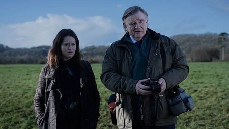 An older man holding a camera and a woman stand together in a grassy field under a cloudy sky with concerned expressions.