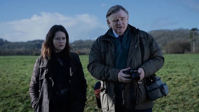An older man holding a camera and a woman stand together in a grassy field under a cloudy sky with concerned expressions.