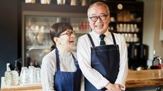 An older man and woman wearing work aprons laugh together at a coffee shop.