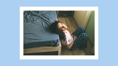 woman sitting on the bedroom floor near her bed and looking sad