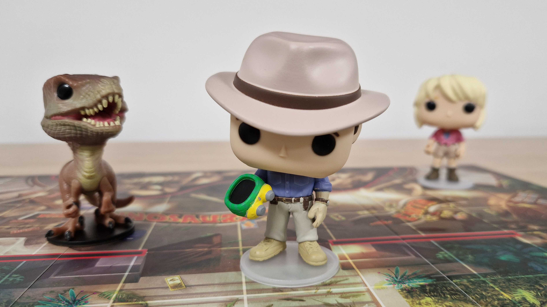 Funko models of a velociraptor, Alan Grant, and Ellie Sattler on the Funkoverse Jurassic Park board