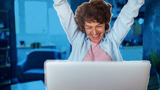 Curly haired woman sitting in front of a laptop at a table, looking very excited with her arms in the air.