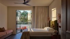 Image of a modern, neutral bedroom with minimalist bedding and a door to a pool area with blackout curtains. There is a fan on the ceiling.
