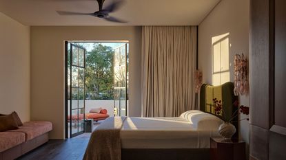 Image of a modern, neutral bedroom with minimalist bedding and a door to a pool area with blackout curtains. There is a fan on the ceiling.