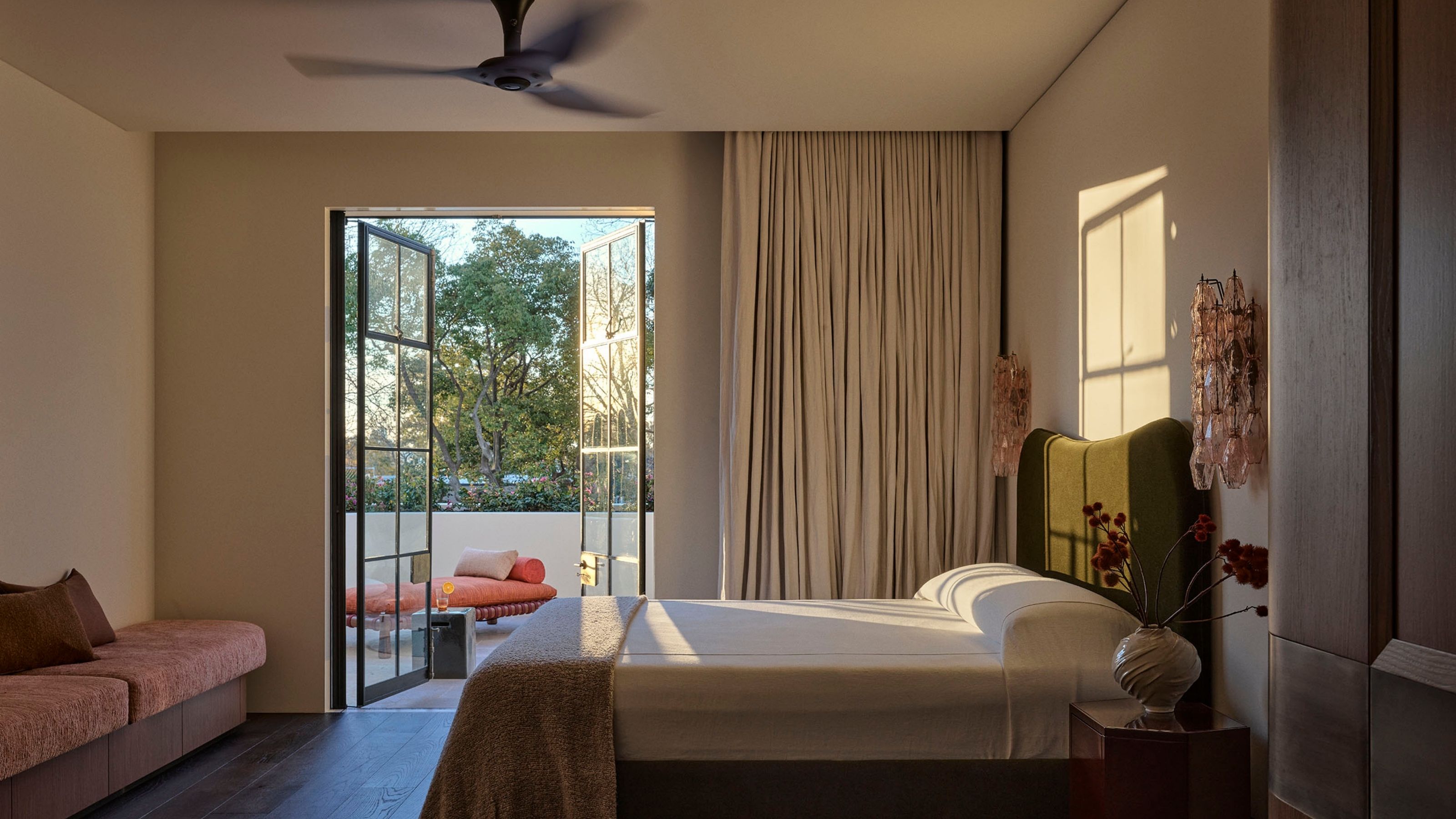 Image of a modern, neutral bedroom with minimalist bedding and a door to a pool area with blackout curtains. There is a fan on the ceiling.