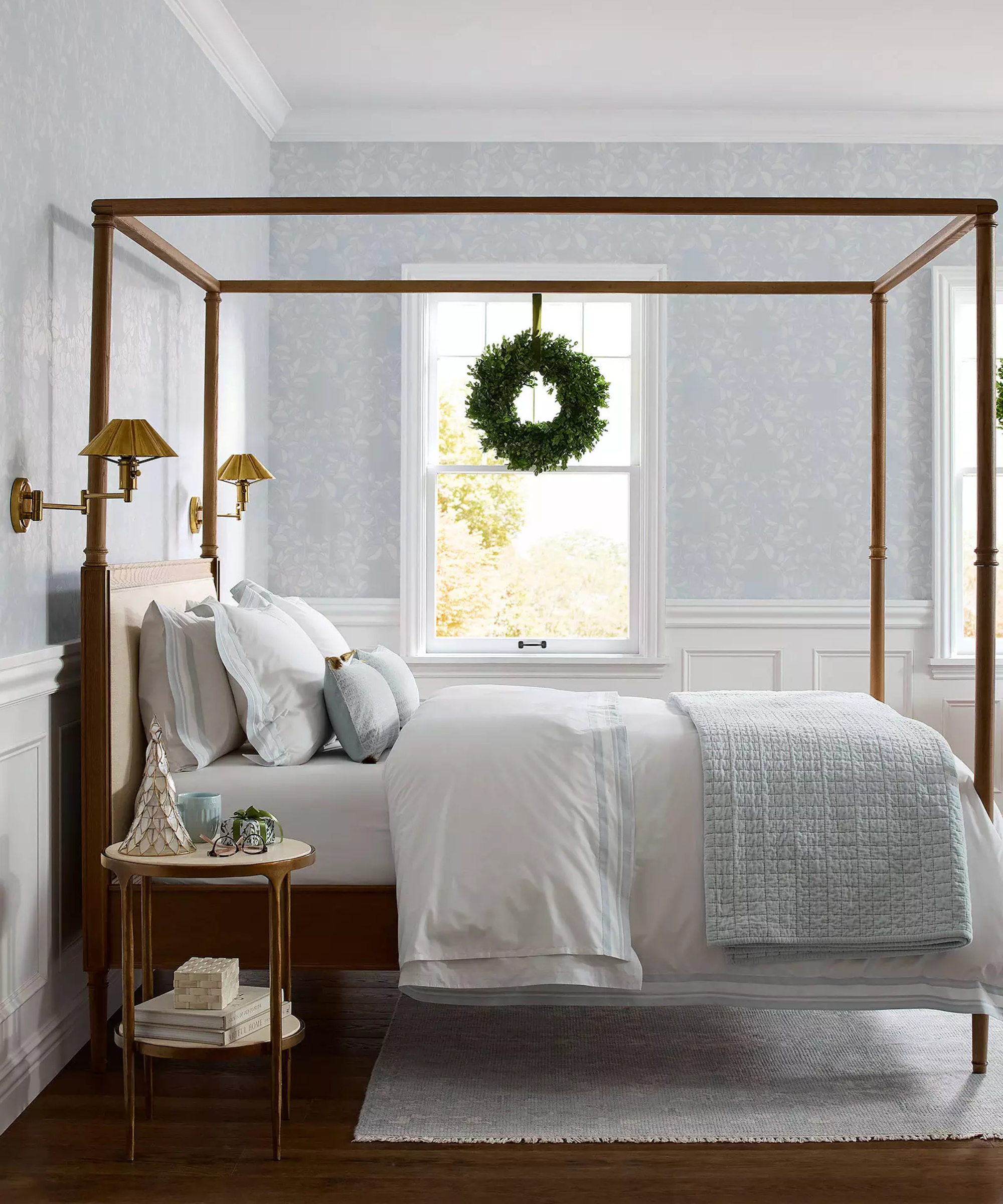 a coastal bedroom with blue wallpaper, a dark wood framed bed, light blue and white sheets, and a wreath hanging from the window