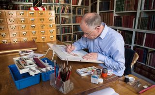 A man in a blue shirt sits over a drawing desk, painting the coat of arms