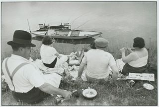 A group of people sit on a grassy river bank with a picnic as a small boat passes by in the background.