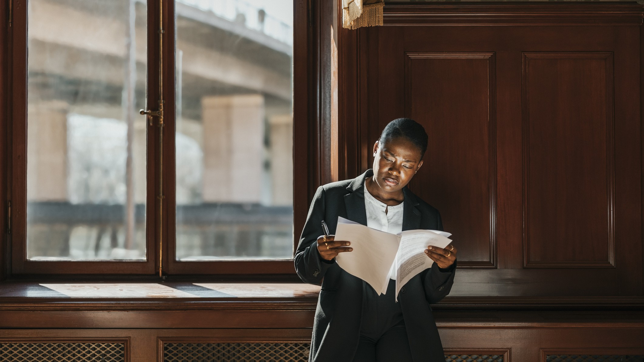 A woman reads a paper document standing in front of a window in a wood-paneled office building.