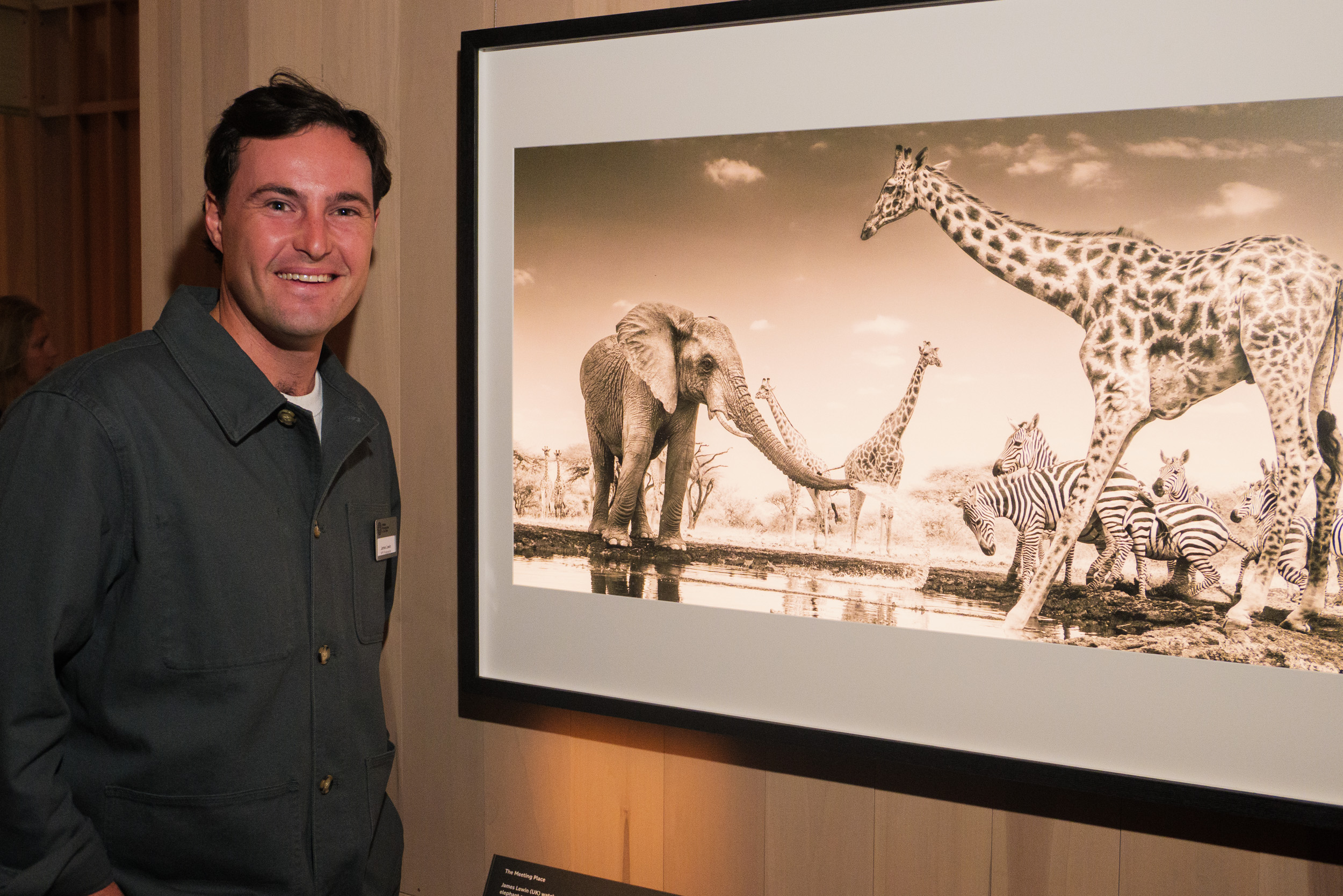 Photographer James Lewin stood next to his winning photograph of an elephant spraying water over zebra and giraffe at a waterhole, at the contest's exhibition in the Natural History Museum, London