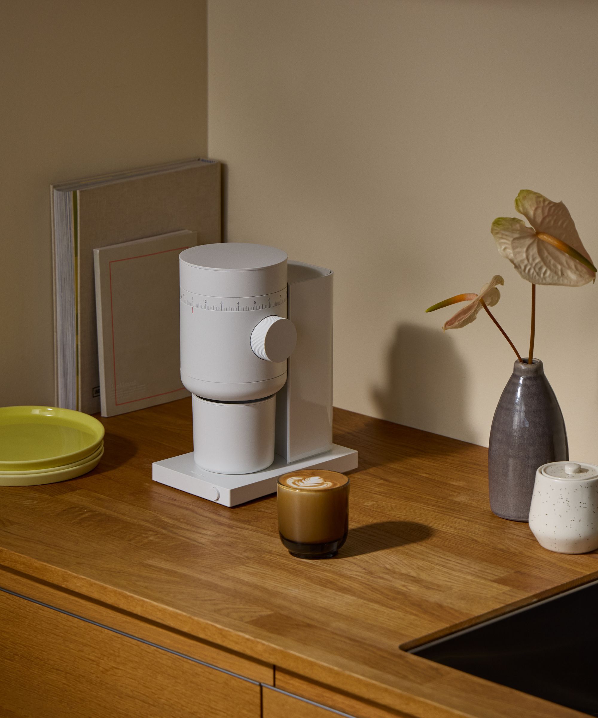 A Fellow Conical Burr Coffee Grinder on a wooden kitchen counter, with a glass cup of coffee beside it.