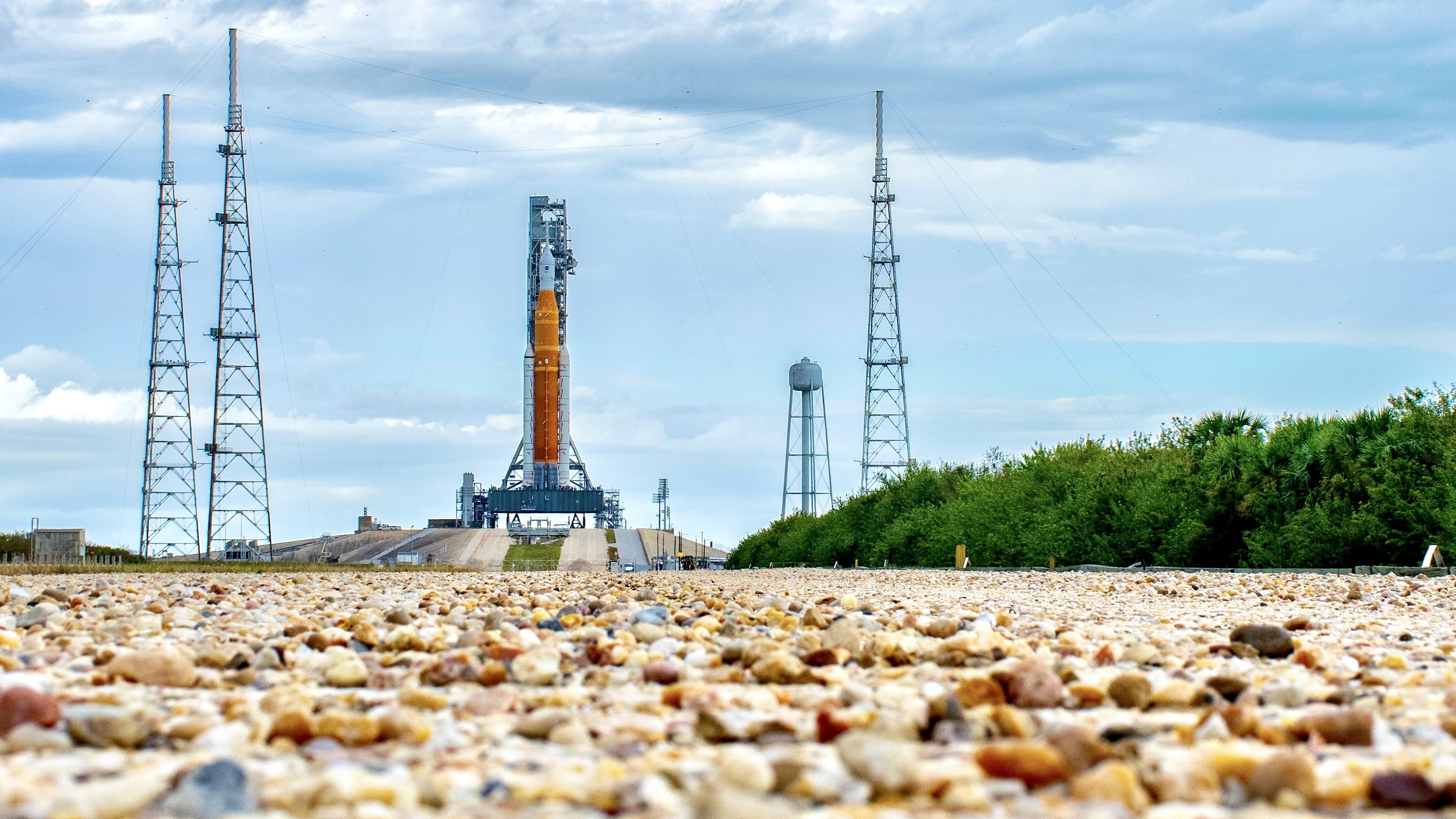 an orange rocket stands in the distance against a blue sky. Rocks lay in the foreground.